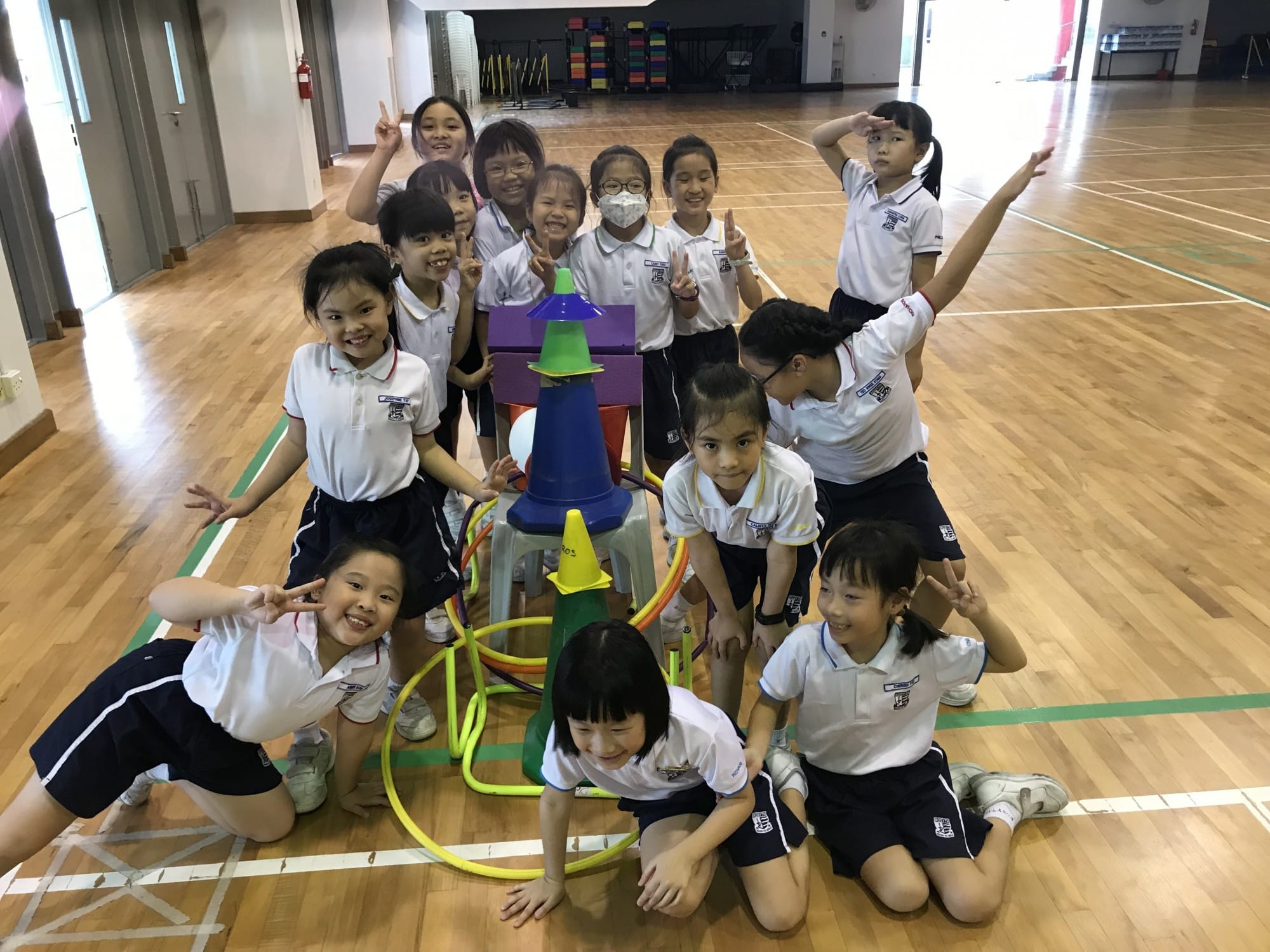A group of young students in white uniforms pose cheerfully in a gym with colorful cones and hoops on a wooden floor.